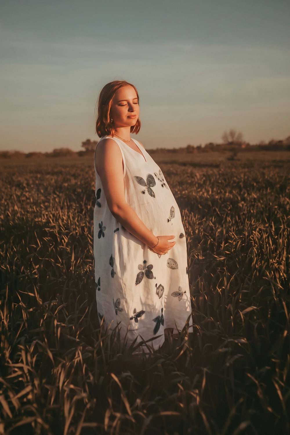 A pregnant woman stands in a field at sunset wearing a sleeveless white dress with black botanical print leaf patterns. The warm golden light illuminates the scene, and the sky is a soft blue-gray.