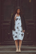 A woman stands on wooden steps in front of a dark carved wooden door, wearing a white midi dress with black botaniocal prints, a black cardigan, and beige flat shoes.
