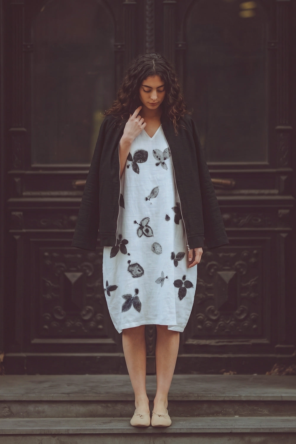 A woman stands on wooden steps in front of a dark carved wooden door, wearing a white midi dress with black botaniocal prints, a black cardigan, and beige flat shoes.