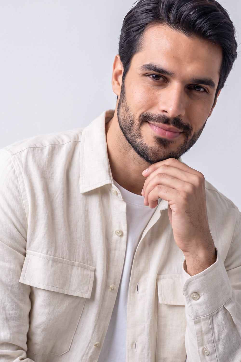 A man with dark hair and a beard wearing a light beige hemp button-up shirt over a white t-shirt, posing against a light gray background.
