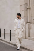 A man wearing a light cream-colored two-piece hemp set consisting of a button-up shirt and matching trousers, styled with a white t-shirt and white sneakers, standing on a city sidewalk next to a stone building.
