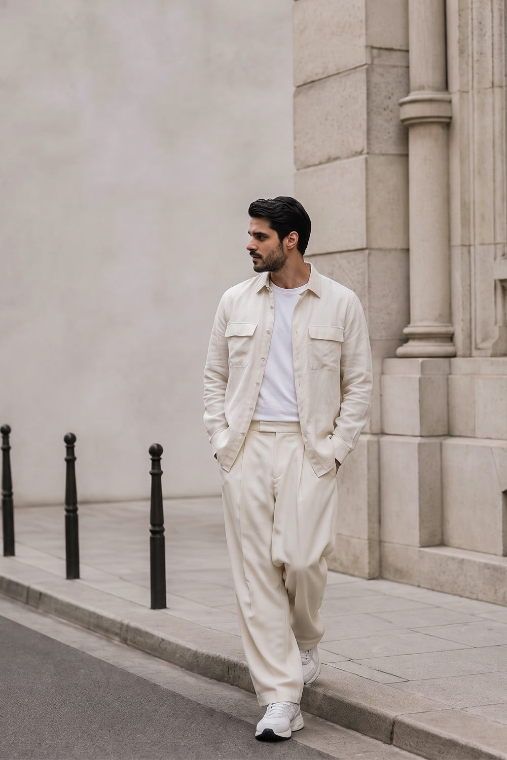 A man wearing a light cream-colored two-piece hemp set consisting of a button-up shirt and matching trousers, styled with a white t-shirt and white sneakers, standing on a city sidewalk next to a stone building.