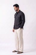 A man wearing a black long-sleeve button-up hemp shirt and off-white linen trousers, standing in profile against a plain white studio background.