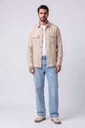 A man modeling a light beige hemp button-up shirt jacket over a white t-shirt and light blue jeans, standing against a light gray studio background.