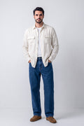 A man wearing a light cream hemp button-up shirt over a white t-shirt, blue jeans, and brown suede shoes, standing against a light gray studio background.