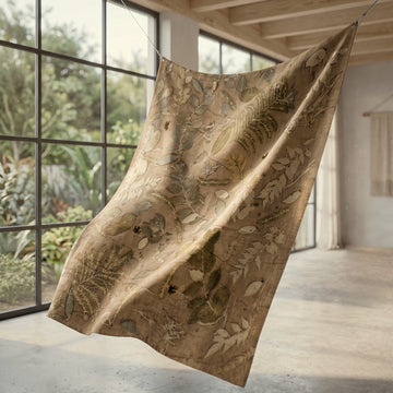 A large beige fabric panel or tapestry featuring a botanical and fossilized leaf pattern, suspended by cords in a modern, sunlit loft interior with large windows.