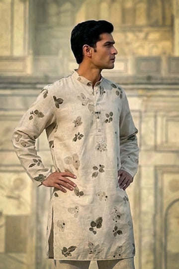 A man wearing a light beige kurta with dark brown floral eco-patterns, posed against a blurred architectural background.