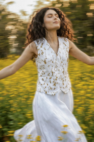Woman twirling in a flower field wearing a sustainable white linen waistcoat with eco-printed golden botanical patterns, paired with a flowing white skirt