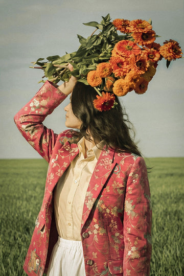 A woman standing in a green field under a pale sky, holding a large bouquet of orange flowers on her head. She is wearing a pink floral blazer over a cream shirt and white skirt.