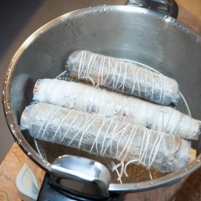 Three cylindrical bundles of fabrics wrapped in white string or fabric, placed inside a large stainless steel pot on a kitchen countertop.