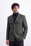 A man wearing an olive green tailored linen blazer with four front flap pockets over a black turtleneck sweater and dark trousers, standing against a plain white studio background.