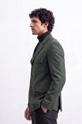 A man in profile wearing an olive green linen blazer over a black turtleneck sweater and dark trousers, standing against a plain white studio background.