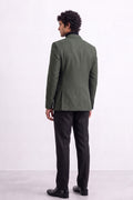 A man viewed from the back wearing an olive green linen blazer, black trousers, and black shoes, standing against a plain white studio background.