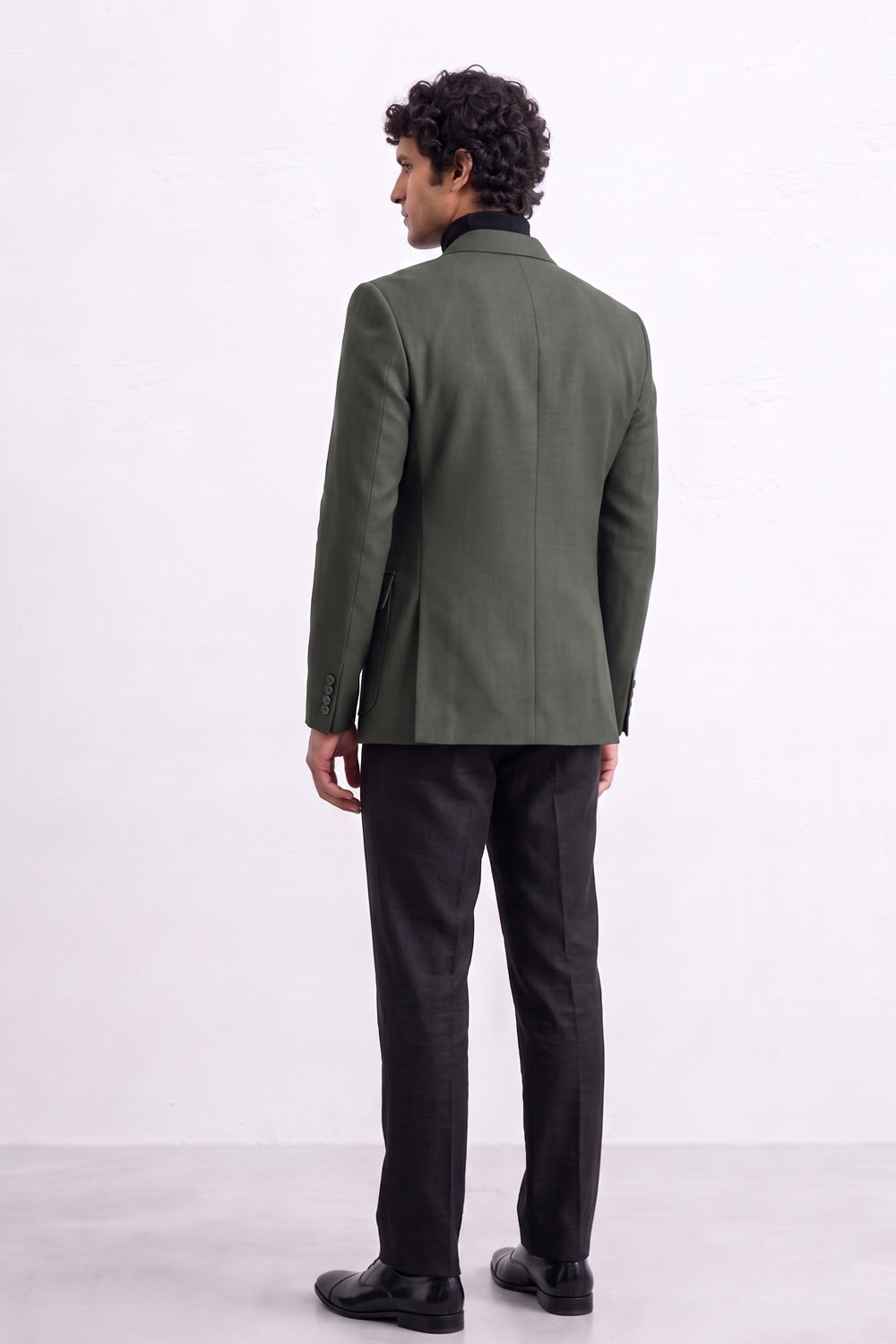A man viewed from the back wearing an olive green linen blazer, black trousers, and black shoes, standing against a plain white studio background.