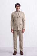 A man modeling a light beige two-piece suit with a matching linen blazer and trousers, worn over a beige turtleneck sweater and paired with brown leather shoes, against a plain light gray studio background.