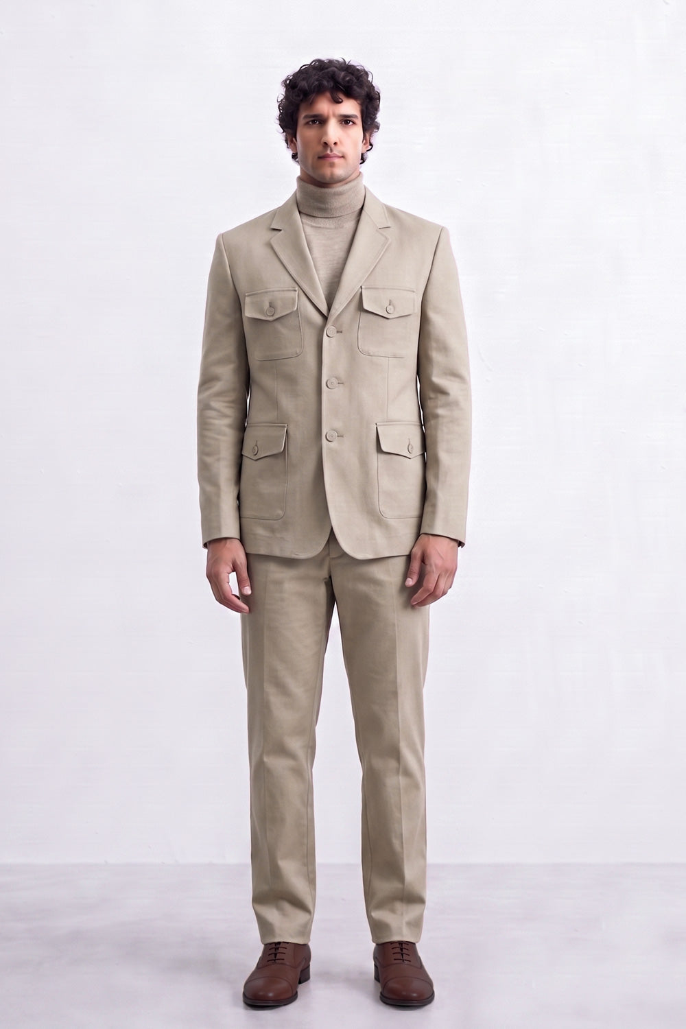 A man modeling a light beige two-piece suit with a matching linen blazer and trousers, worn over a beige turtleneck sweater and paired with brown leather shoes, against a plain light gray studio background.
