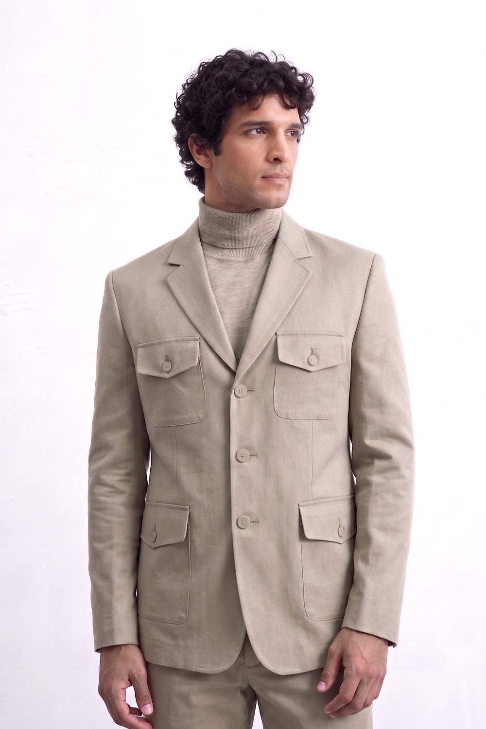 A man wearing a light beige linen-blazer with four front flap pockets over a matching beige turtleneck sweater and trousers, photographed against a plain white studio background.