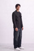 A man wearing a black linen blazer over a black t-shirt, dark gray jeans, and white sneakers, standing in profile against a light gray studio background.
