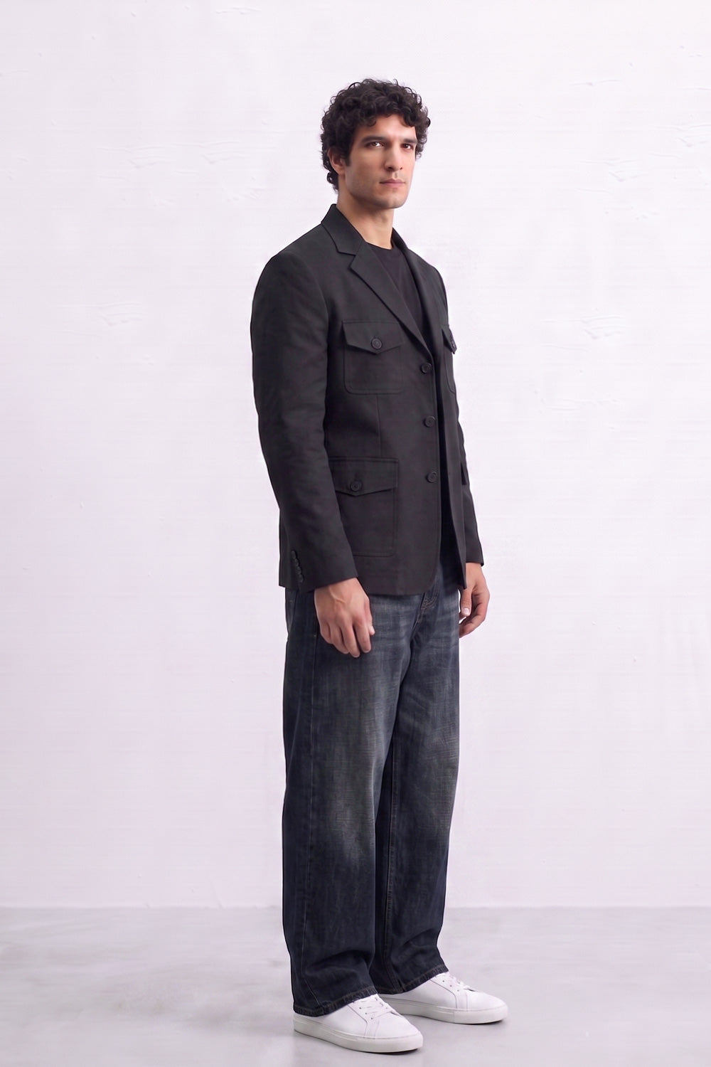 A man wearing a black linen blazer over a black t-shirt, dark gray jeans, and white sneakers, standing in profile against a light gray studio background.