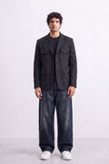 A man stands against a light gray studio background wearing a dark charcoal gray linen blazer over a black shirt, loose dark blue jeans, and white sneakers.