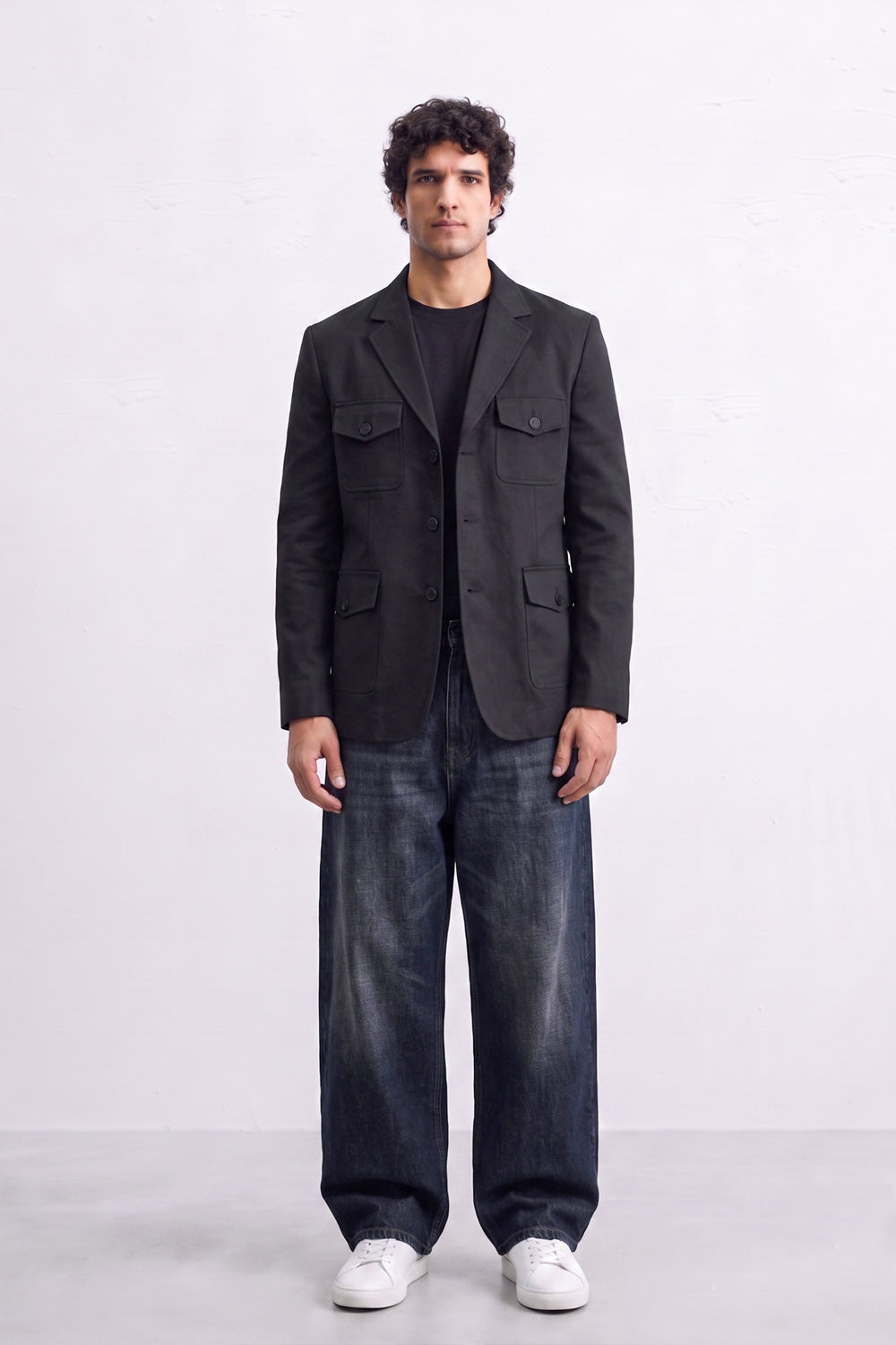 A man stands against a light gray studio background wearing a dark charcoal gray linen blazer over a black shirt, loose dark blue jeans, and white sneakers.