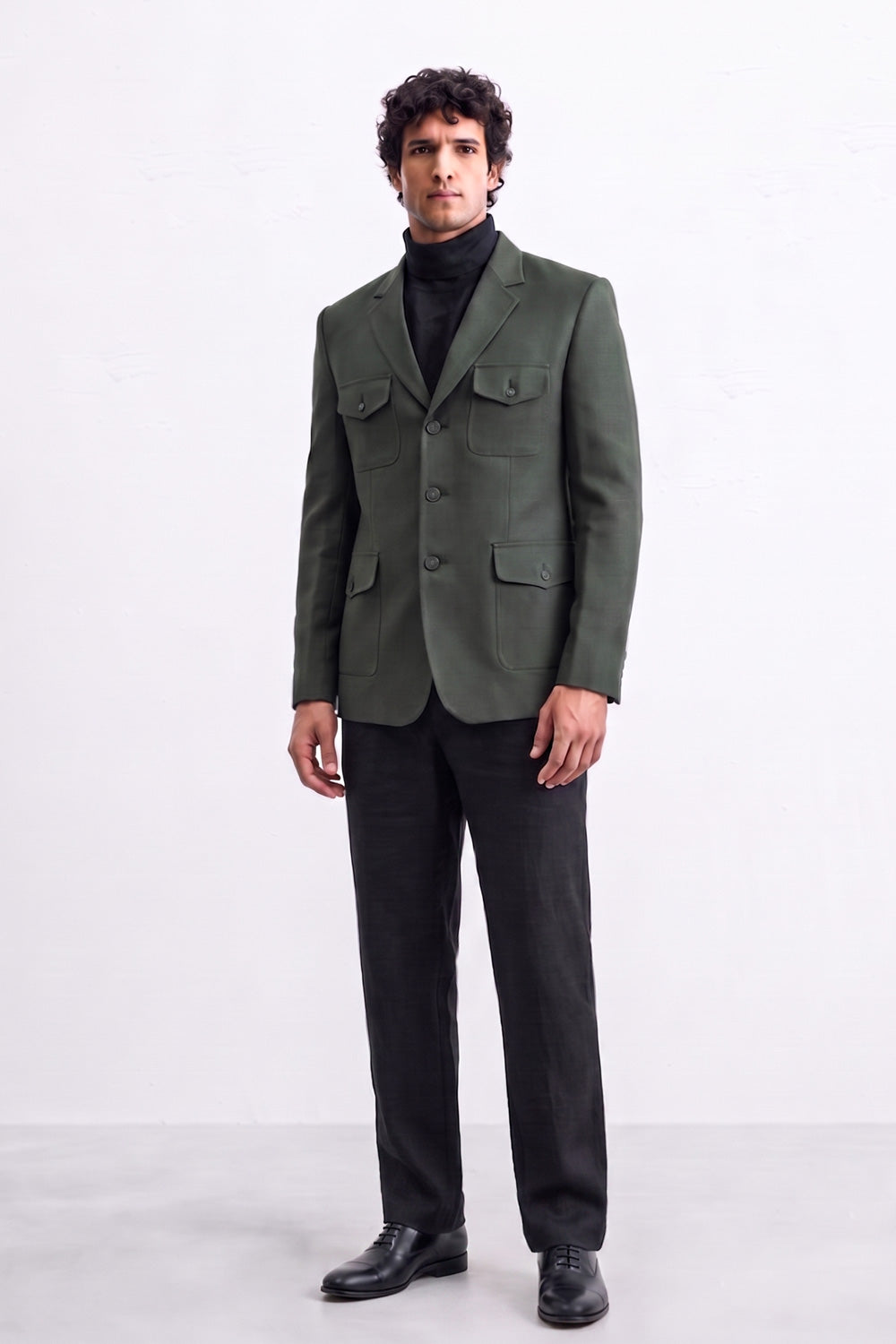 A man wearing a dark olive green linen blazer with flap pockets over a black turtleneck sweater, black trousers, and black leather shoes, standing against a plain white studio background.
