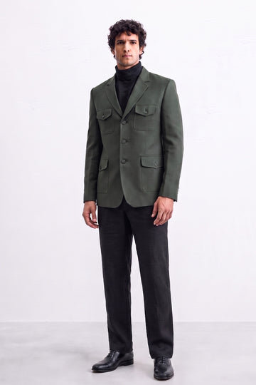 A man wearing a dark olive green linen blazer with flap pockets over a black turtleneck sweater, black trousers, and black leather shoes, standing against a plain white studio background.