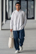 A man walking in a modern indoor corridor wearing a white long-sleeve linen shirt, loose dark blue jeans, and white sneakers, while carrying a beige canvas tote bag.