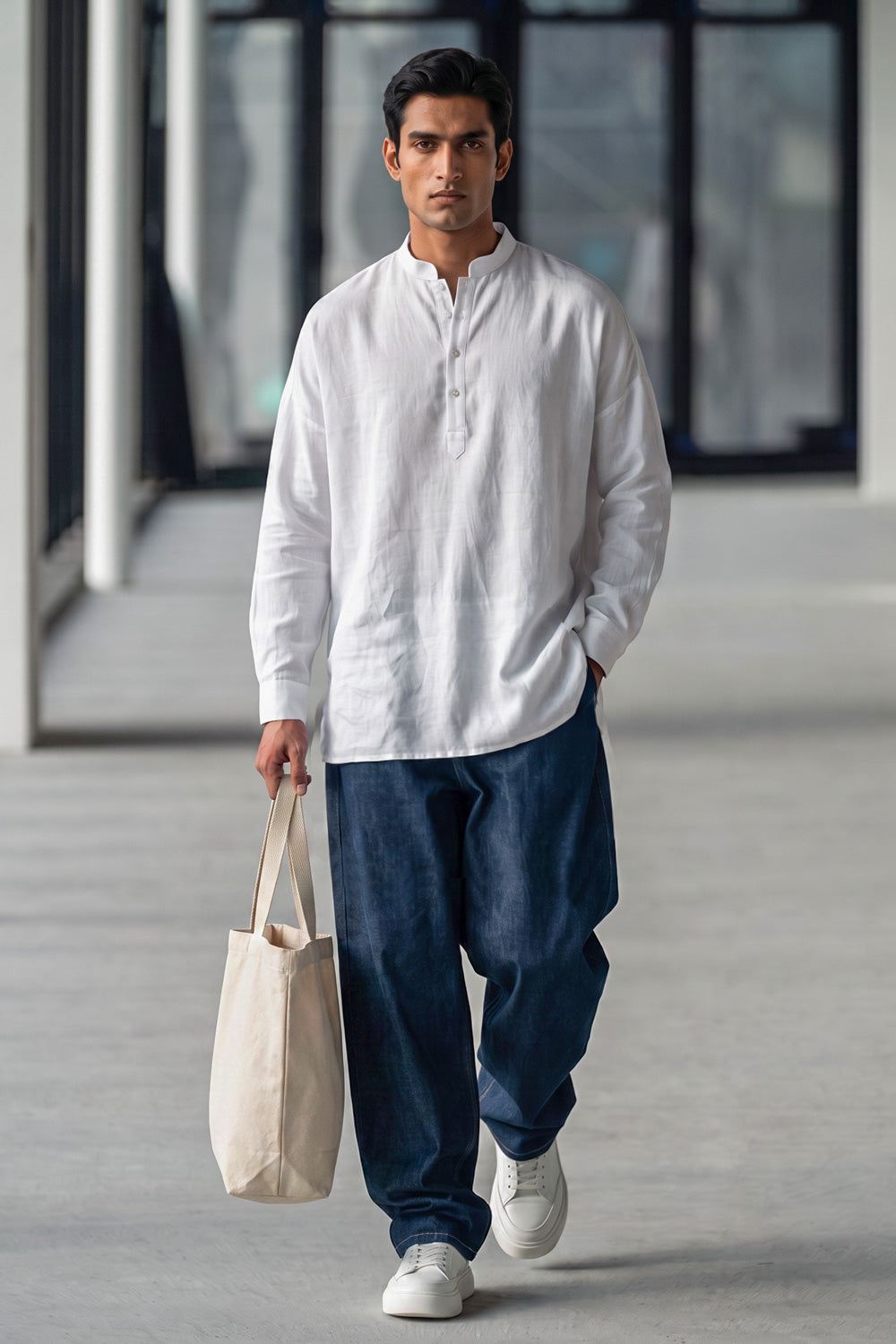 A man walking in a modern indoor corridor wearing a white long-sleeve linen shirt, loose dark blue jeans, and white sneakers, while carrying a beige canvas tote bag.