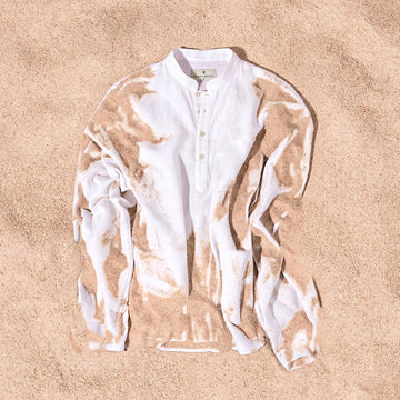 A white long-sleeve henley shirt with a sand-dyed pattern, laid flat on a bed of beige sand.