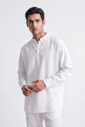 A man wearing a white long-sleeve linen kurta with a mandarin collar and button placket, paired with matching white trousers, photographed against a seamless light gray studio background.