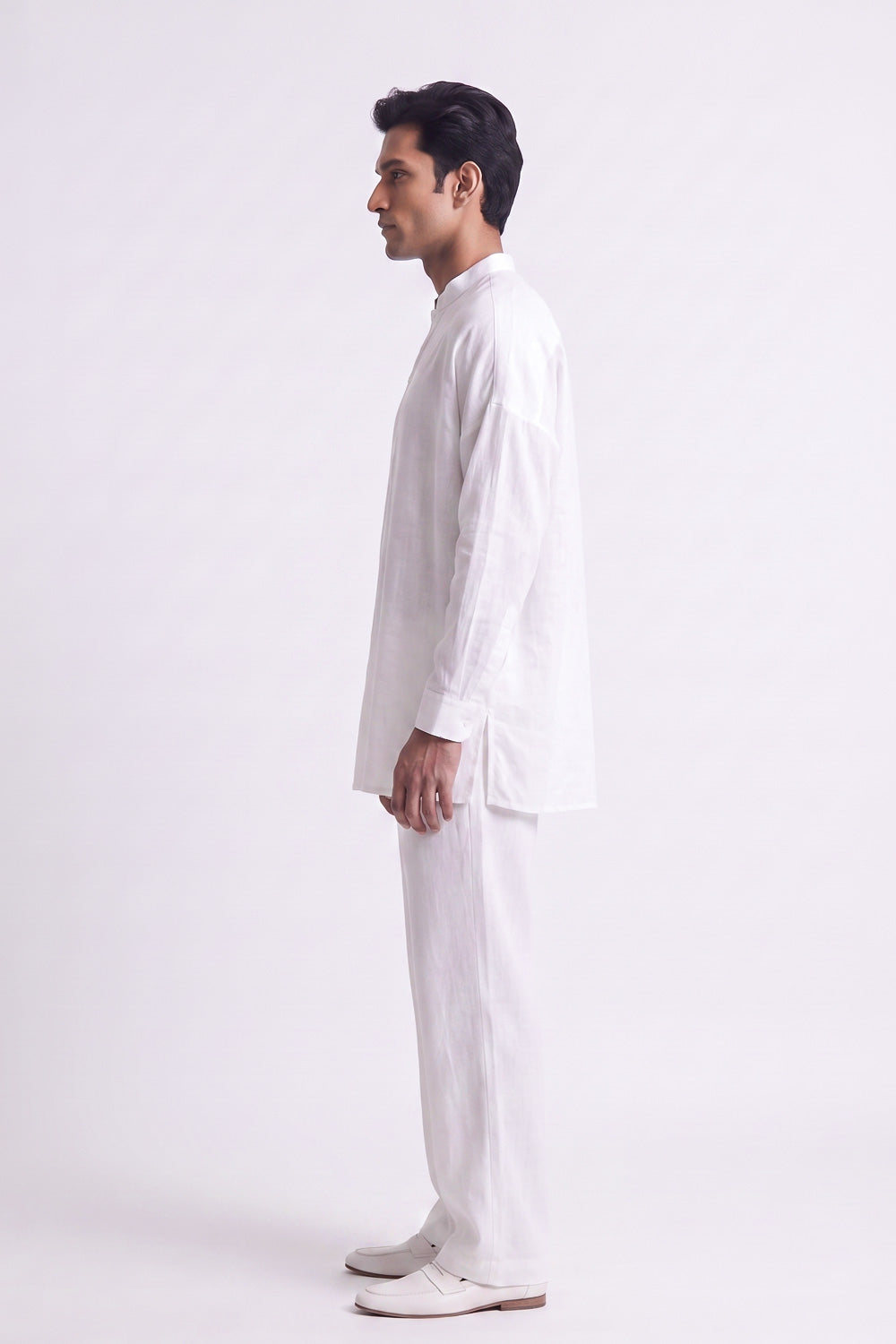A man in a white long-sleeve linen kurta and matching trousers, standing in profile against a plain white studio background.