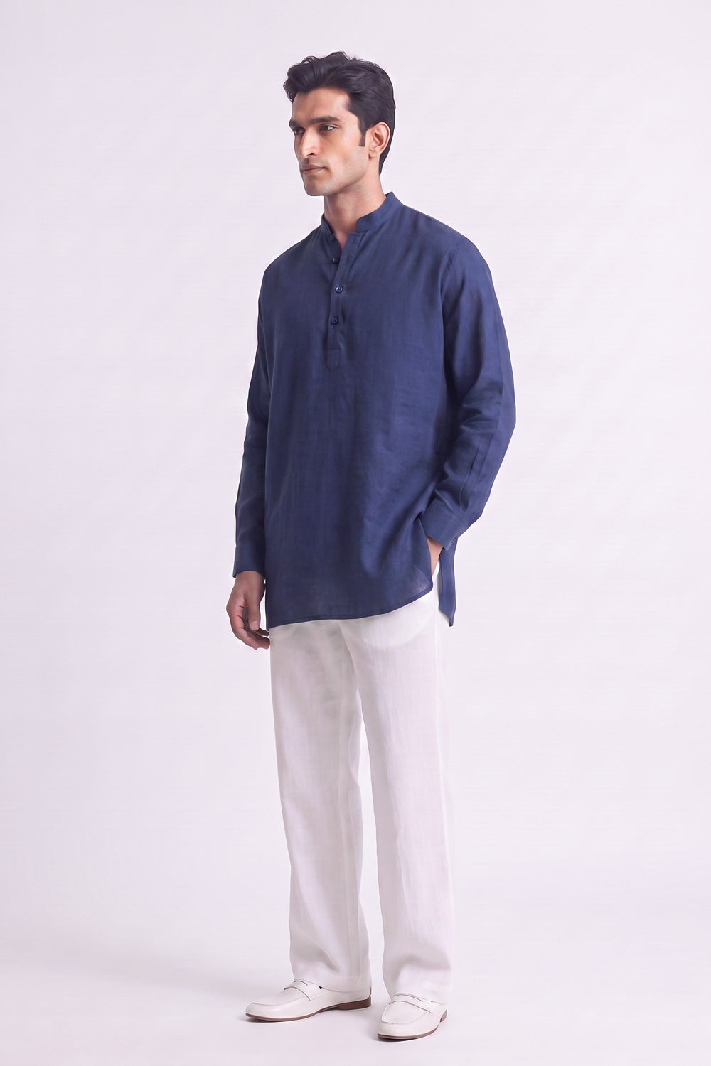 A man modeling a navy blue long-sleeve linen kurta with a mandarin collar and three-button placket, paired with off-white trousers and white shoes, against a plain light gray studio background.