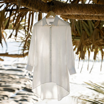A white long-sleeve linen button-up linen  shirt hangs from a wooden hanger on a tree branch outdoors, with a beach and palm leaves blurred in the background.