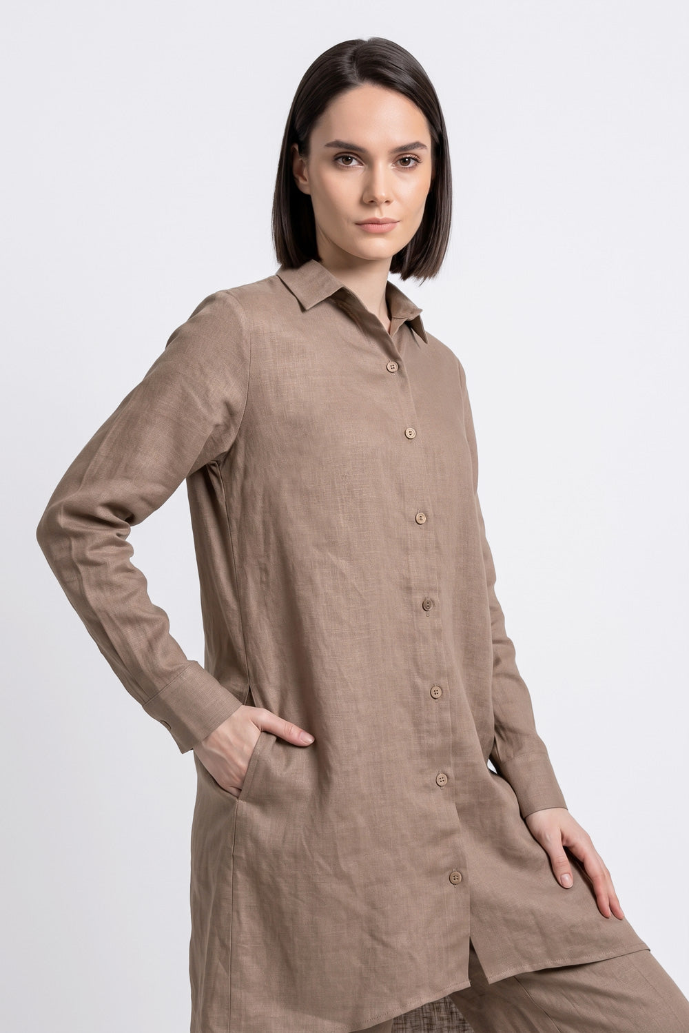 A woman modeling a long-sleeve, button-front linen shirt dress in a warm taupe-brown color, paired with matching trousers, against a plain light gray studio background.