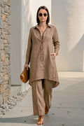 A woman walking outdoors wearing a taupe-brown linen long-sleeve button-up shirt and matching wide-leg trousers, accessorized with sunglasses, layered necklaces, and a brown clutch.