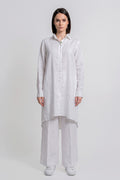 A woman stands against a light gray studio background wearing a loose white long-sleeve button-front linen  shirt with wide-leg white pants and white sneakers.