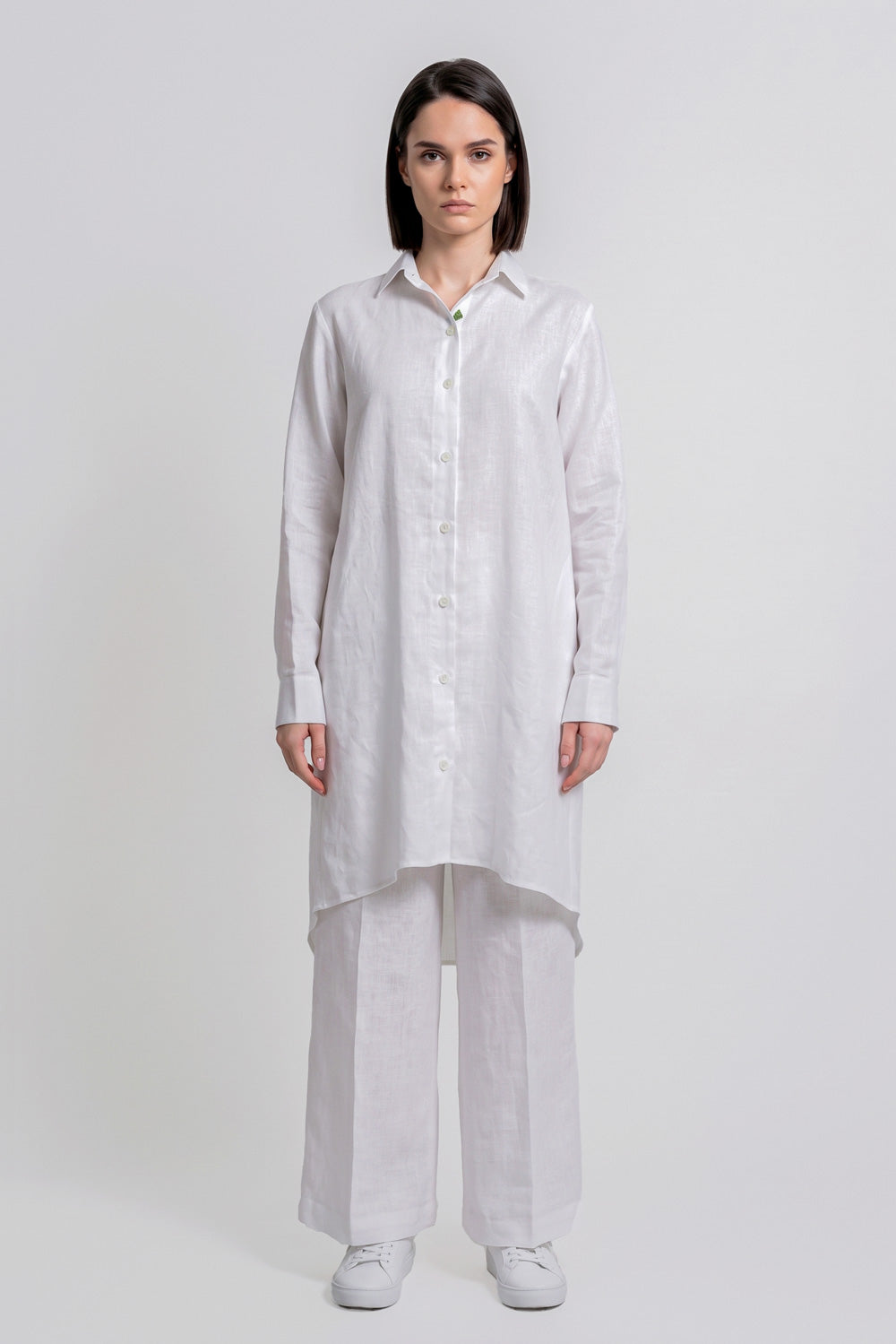 A woman stands against a light gray studio background wearing a loose white long-sleeve button-front linen  shirt with wide-leg white pants and white sneakers.