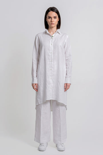 A woman stands against a light gray studio background wearing a loose white long-sleeve button-front linen  shirt with wide-leg white pants and white sneakers.