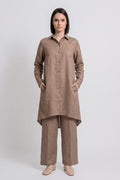 A woman stands against a light gray studio background wearing a loose brown long-sleeve button-front linen  shirt with wide-leg white pants and beige shoes.