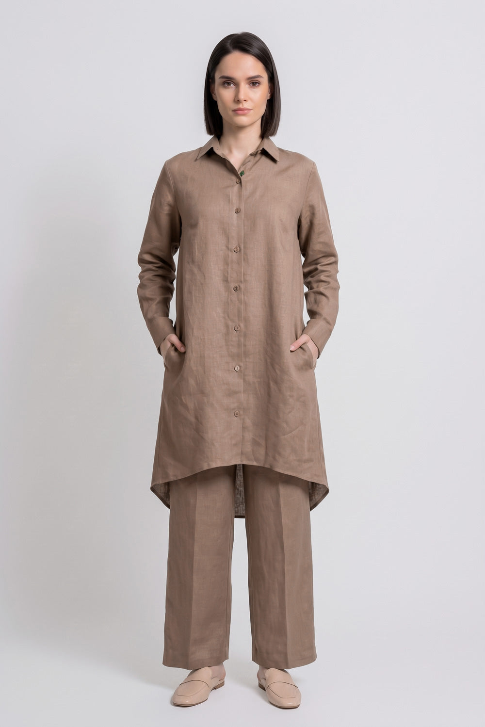 A woman stands against a light gray studio background wearing a loose brown long-sleeve button-front linen  shirt with wide-leg white pants and beige shoes.