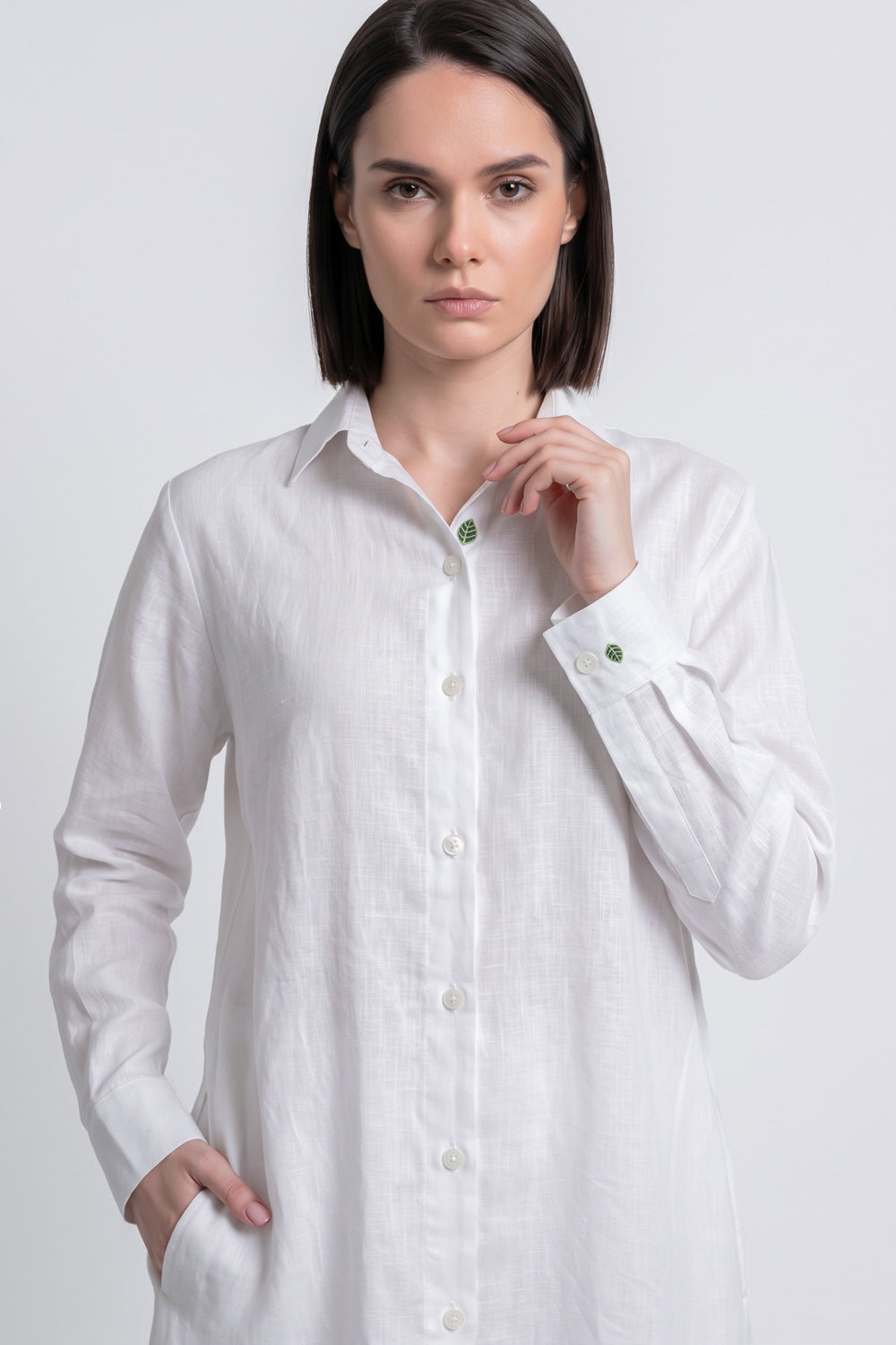 A woman modeling a white long-sleeve linen shirt with green buttons, set against a plain light gray studio background.