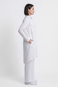 A woman in a white long-sleeve button-up linen shirt and matching white trousers, shown in profile against a plain light gray studio background.