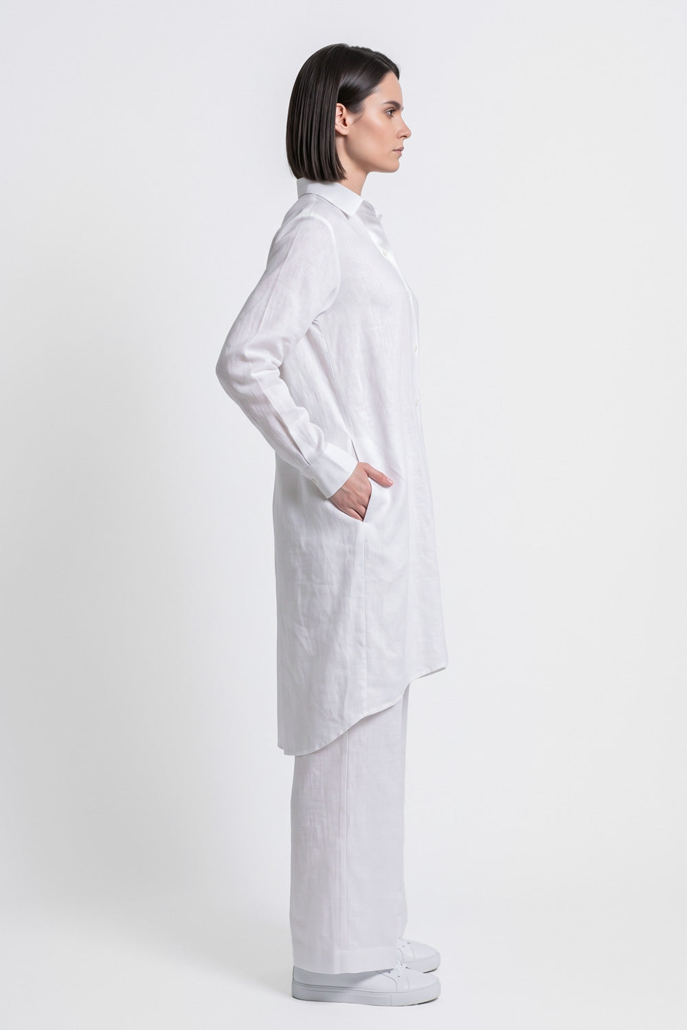 A woman in a white long-sleeve button-up linen shirt and matching white trousers, shown in profile against a plain light gray studio background.