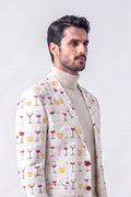 A man wearing a cream-colored blazer with a repeating pattern of wine glasses filled with red, yellow, and white liquids, layered over a beige turtleneck sweater, set against a plain light gray background.