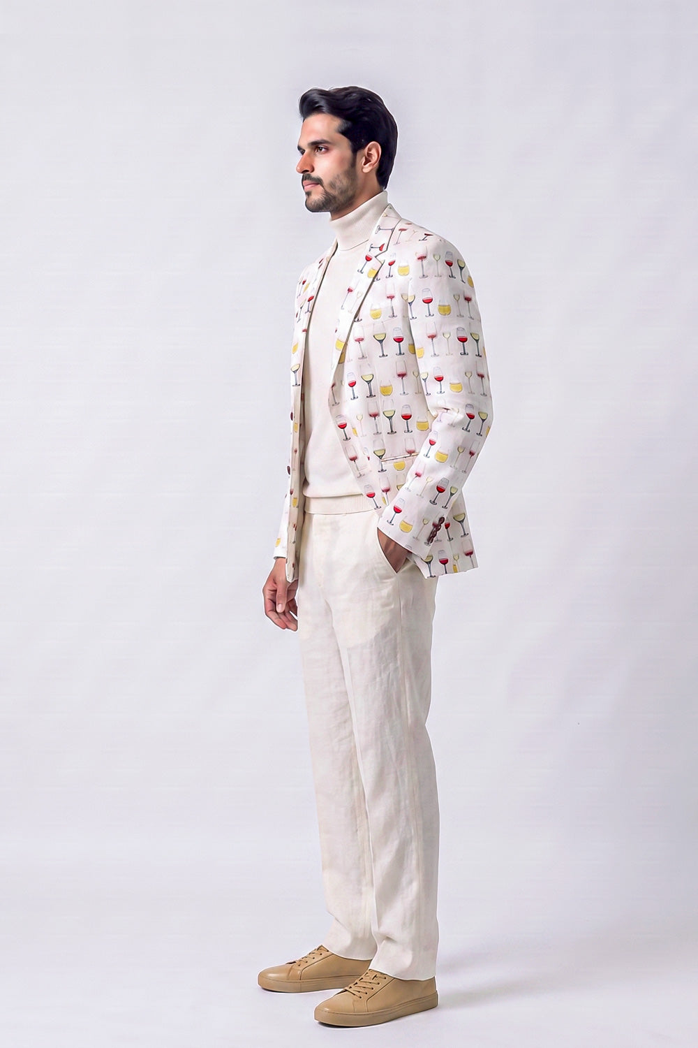 A man wearing a cream-colored blazer with a colorful cocktail print, a cream turtleneck sweater, cream trousers, and beige sneakers, standing in profile against a light gray studio background.