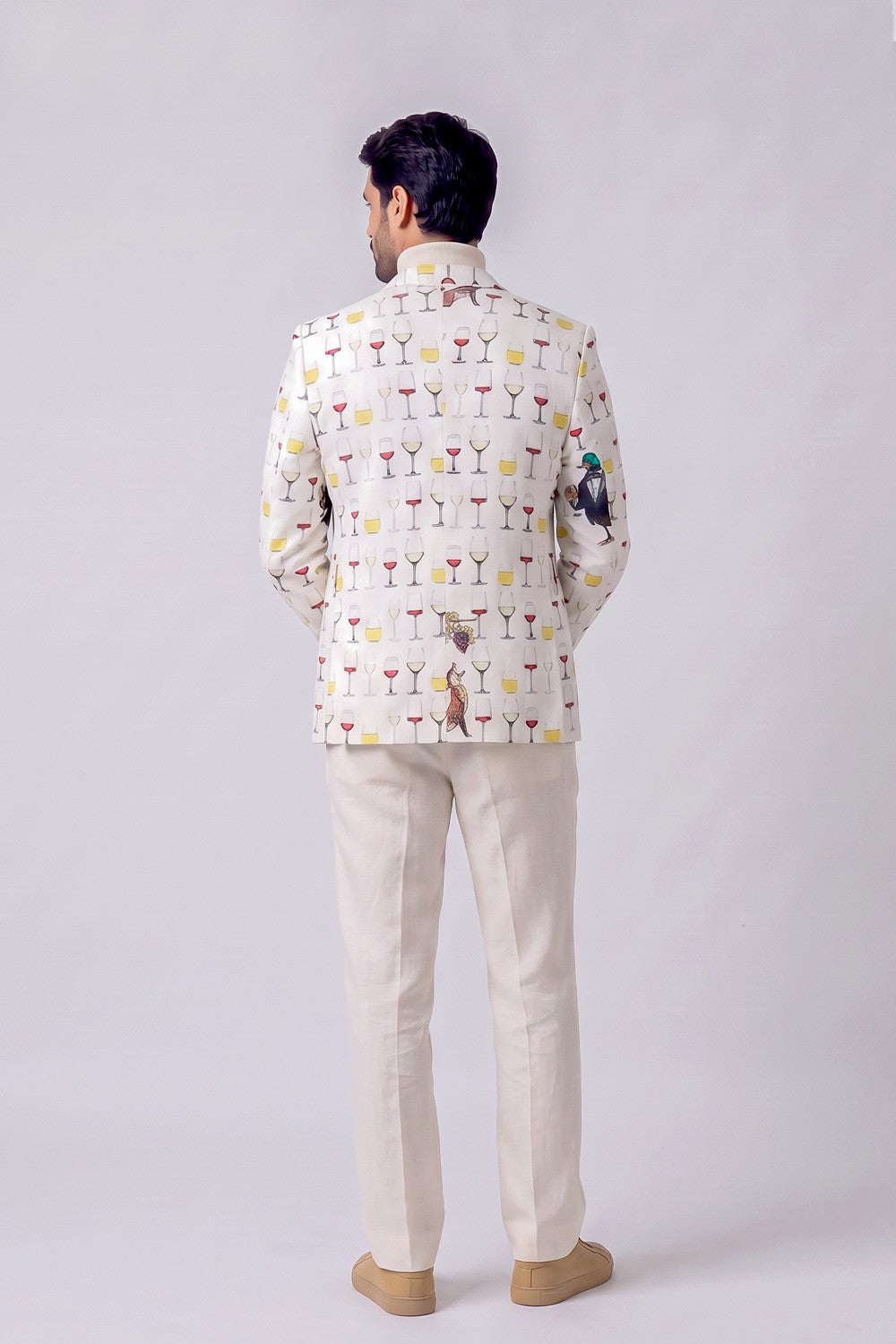 A man viewed from the back wearing a cream-colored Nehru jacket with a colorful cocktail glass pattern and beige trousers, standing against a light gray studio background.