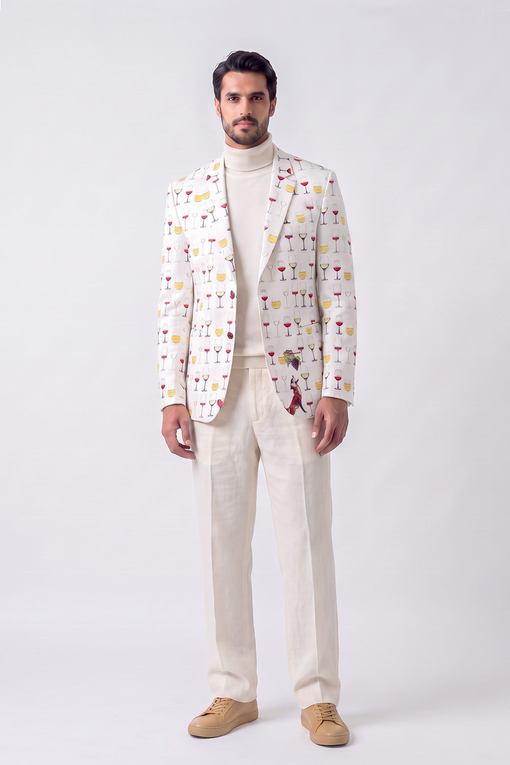 A man modeling a cream-colored blazer with a colorful cocktail glass pattern, paired with a cream turtleneck, cream trousers, and tan suede loafers, against a light gray studio background.