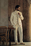 A man wearing a cream-colored suit with a colorful cocktail glass pattern on the blazer, paired with a cream turtleneck and cream trousers, standing in a rustic room with wooden walls.