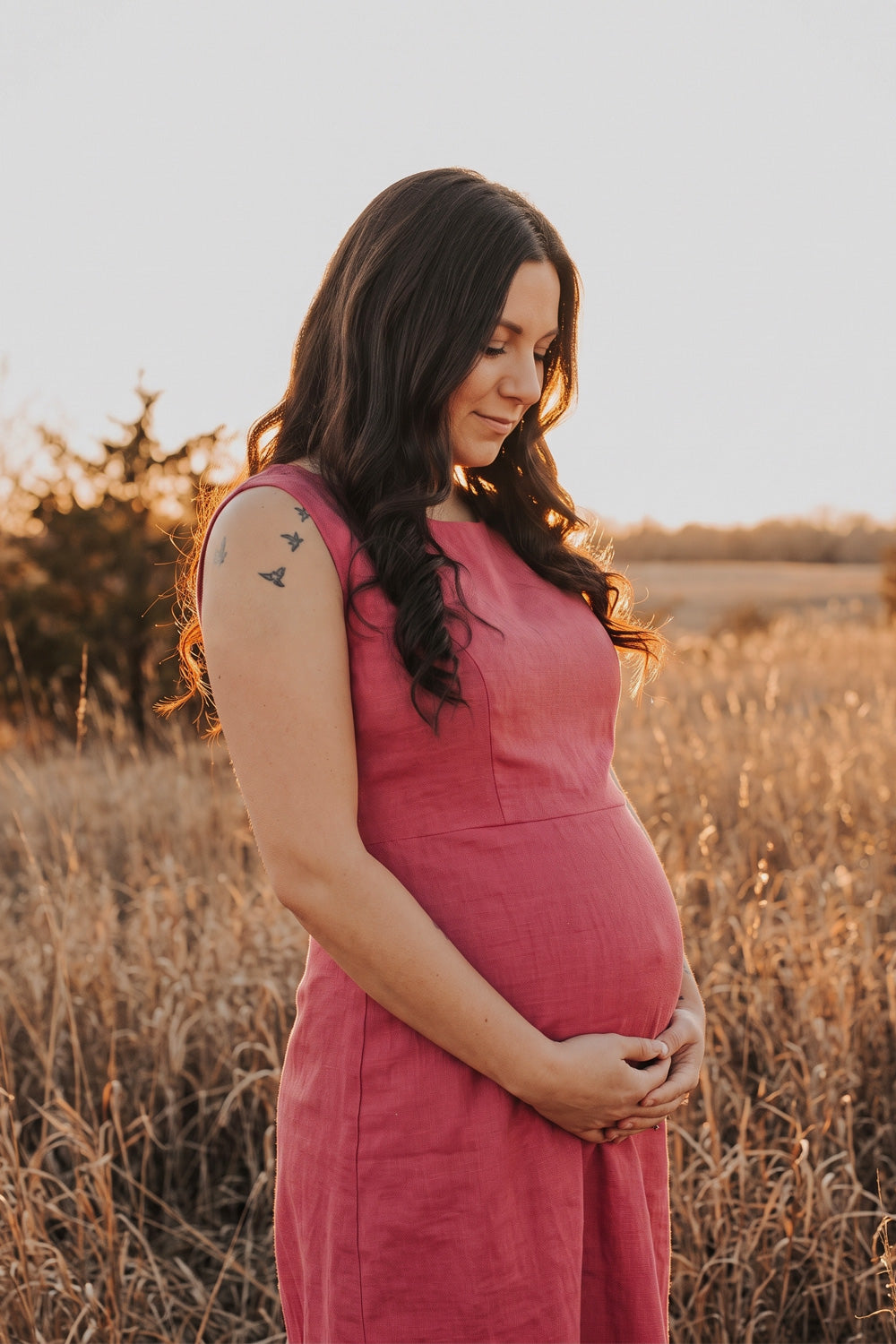 A pregnant woman wearing a sleeveless dusty rose pink linen dress stands in a field of dry golden grass at sunset, cradling her baby bump.